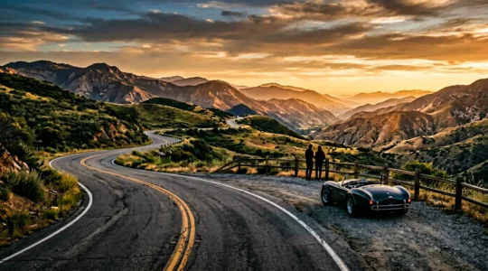Scenic road winding through mountains during golden hour with a vintage car parked at an overlook
