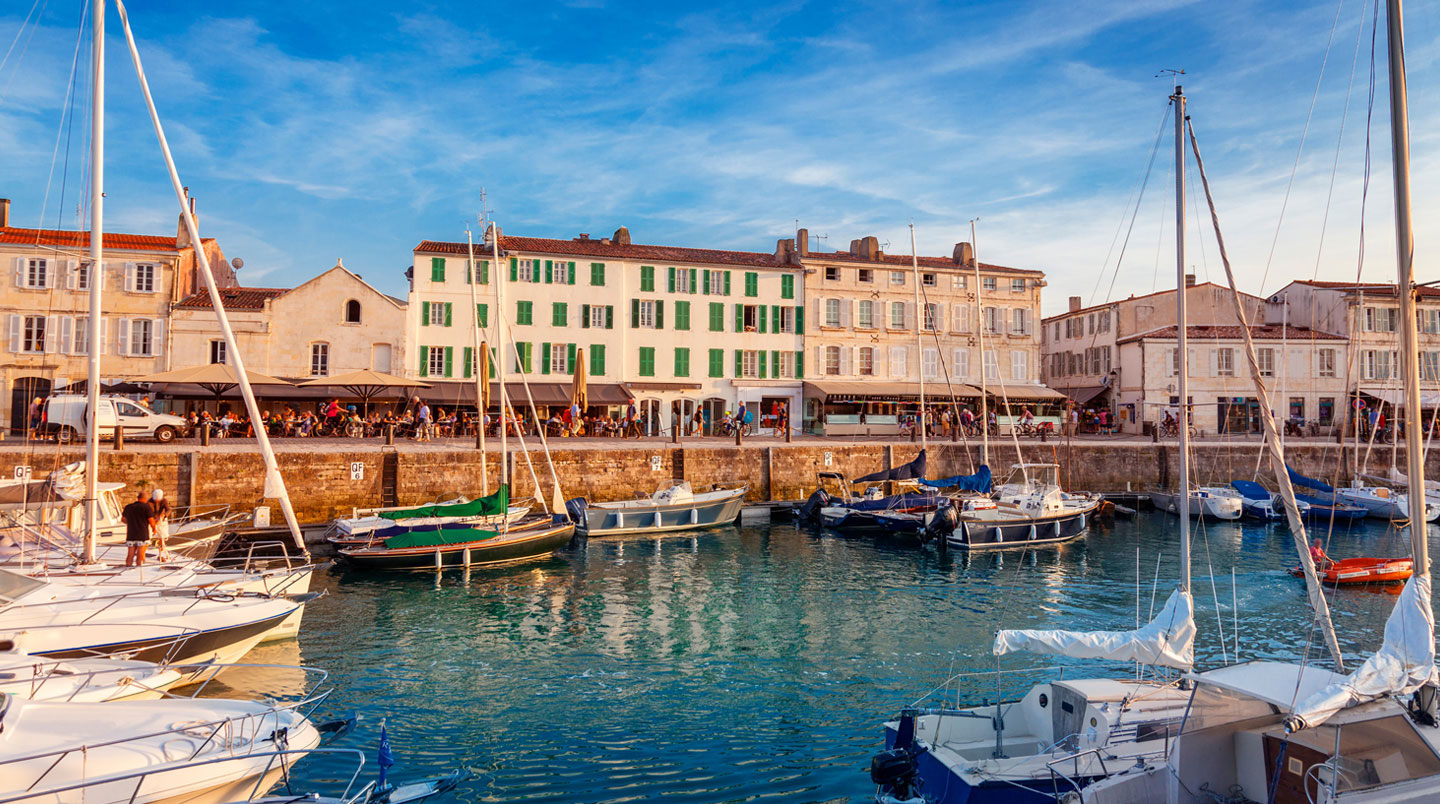 People walking along a harbour at dusk with café terraces lit and boats moored nearby.