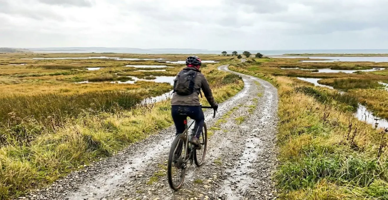 A cyclist seen from behind pedalling along a coastal path through marshlands, with salt flats and open sky visible in the distance