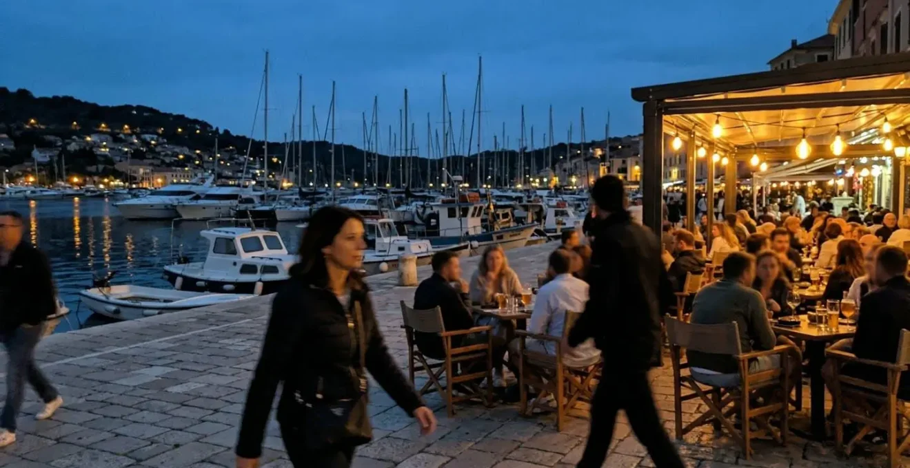 People walking along a harbour at dusk with café terraces lit and boats moored nearby.