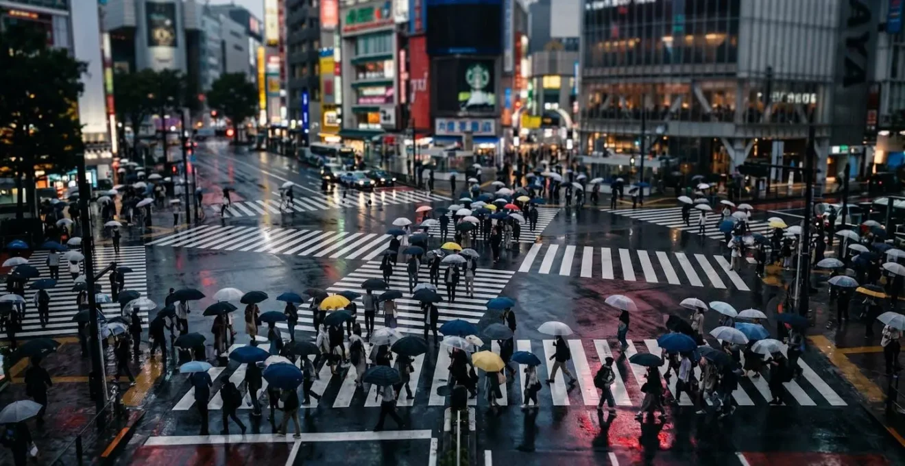 Aerial view of Shibuya crossing on a rainy night with colorful umbrellas creating patterns on the wet, reflective pavement.